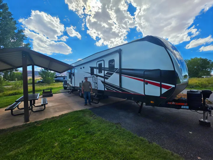 A man stands by a large, white and black fifth-wheel rv rental at a sunny campsite with a picnic table.. Cruiser Rv Corp Stryker 2025