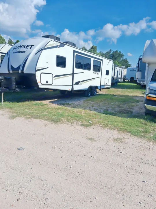 White Sunset Super Lite travel trailer RV rental, prominently parked in an outdoor lot. Other campers visible under a blue sky.. Crossroads RV Sunset Trail 2022