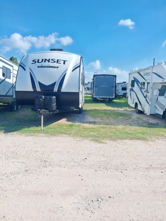 RV rental lot featuring a white 'Sunset Super Lite' travel trailer, motorhomes, and other campers parked on gravel under a blue sky.. Crossroads RV Sunset Trail 2022