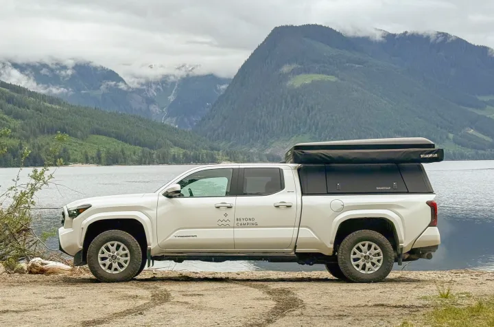 Overlanding rv rental: White Toyota Tacoma pickup with SmartCap camper & rooftop tent by a scenic mountain lake under cloudy skies.. Toyota Tacoma 2020
