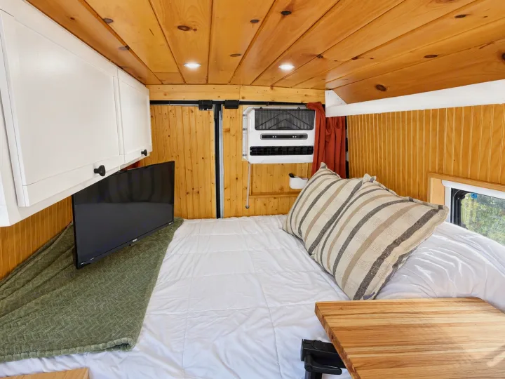 Cozy RV rental interior: a comfy bed, TV, AC unit, white cabinets, & warm wood paneling furnish this inviting camper van.. Dodge Ram 2500 2024