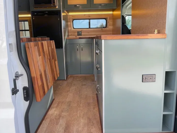 Modern rv rental van conversion kitchen with light green cabinets, wood counters, & fold-out table. Cork floor.. Off Grid Adventure Vans Sarge 2019
