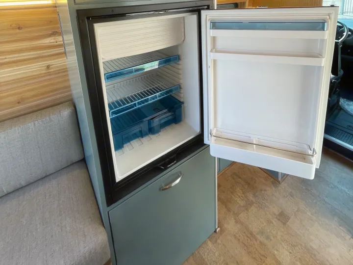 RV rental: Open refrigerator revealing compact interior, shelves, and freezer compartment, beside a cozy seating area.. Off Grid Adventure Vans Sarge 2019