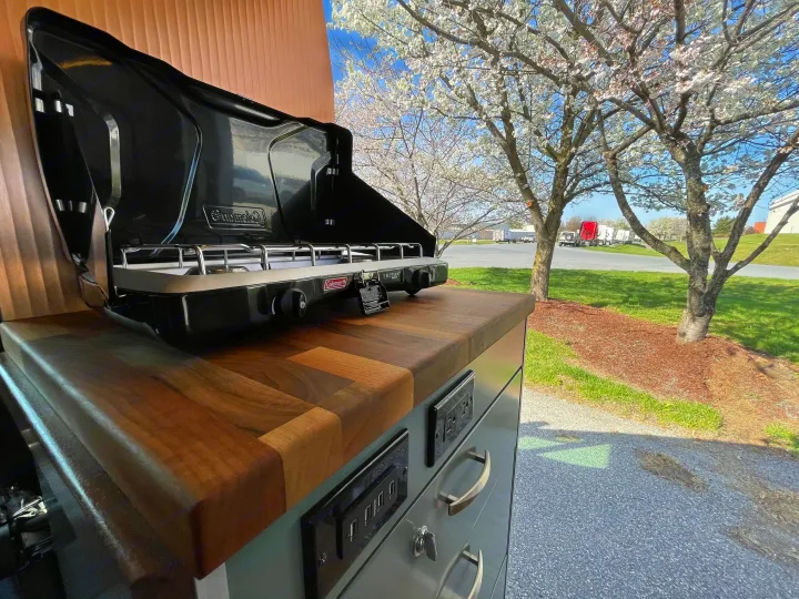 Inside an RV rental: a Coleman Triton camping stove on a wood counter, with blooming trees and a sunny sky outside.. Off Grid Adventure Vans Sarge 2019