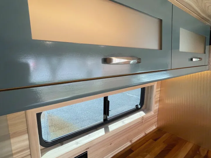 RV rental interior features blue-green overhead cabinets, a window with wood trim, and warm lighting on wood paneling.. Off Grid Adventure Vans Sarge 2019