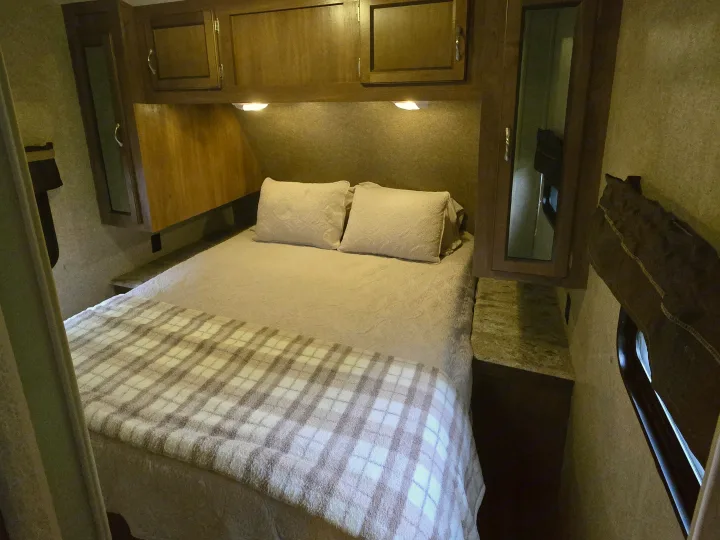 Cozy RV rental bedroom with warm lighting, overhead cabinets, and a comfortable bed with pillows and a plaid blanket.. Coachmen Catalina 2017