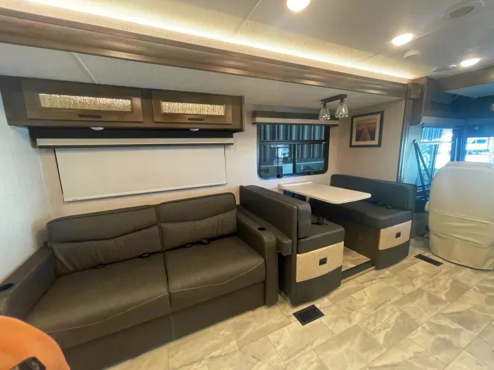 Modern RV rental interior: dark sofa, dinette with table, overhead cabinets, and light tiled floor for comfortable travel.. Coachmen MIRADA 35ESF 2026