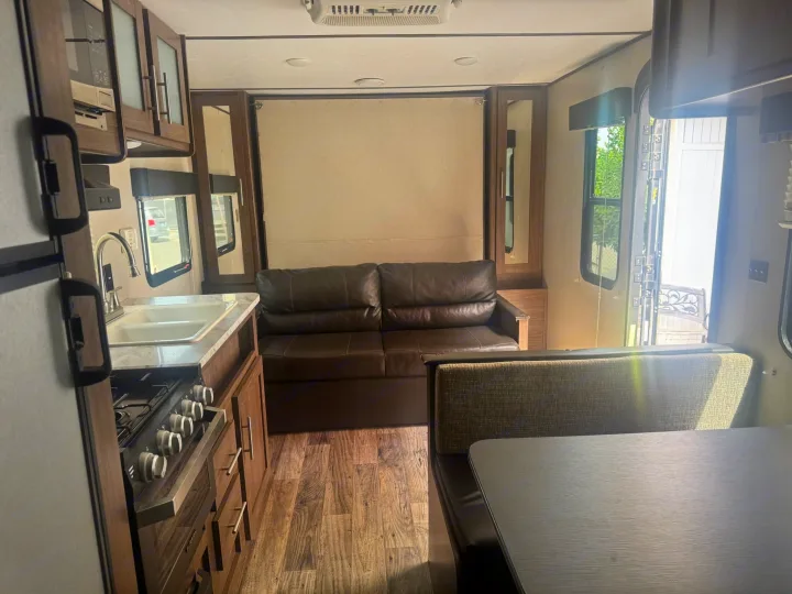 Interior of an RV rental: kitchen with stove & sink, dinette seating, and a comfortable brown sofa on wood-look flooring.. Keystone RV Hideout LHS 2019