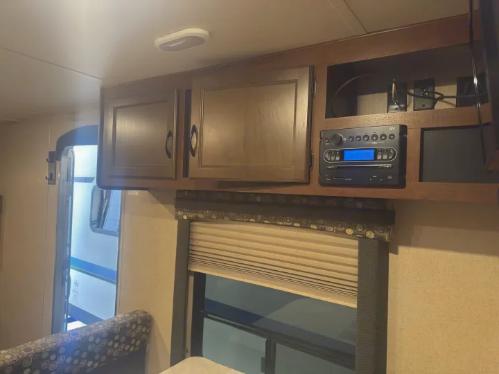 Inside an **rv rental**, showing wooden overhead cabinets, an iRV stereo, and a window with pleated blinds.. Jayco Jay Flight 2015