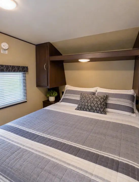 This cozy RV rental bedroom offers a comfortable bed with striped linens and convenient overhead storage.. Keystone RV Springdale SSR 2018