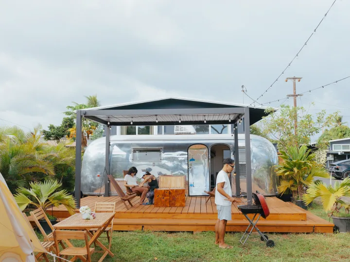 A family enjoys their Airstream rv rental, grilling and relaxing on a wooden deck amidst tropical greenery. Perfect outdoor living.. Airstream Land Yacht 1960