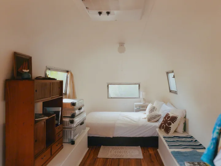 This bright RV rental bedroom features a cozy bed with white linens, wooden furnishings, and windows looking out to tropical greenery.. Airstream Land Yacht 1960