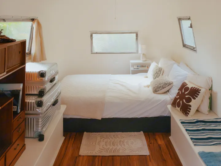 Cozy, minimalist RV rental bedroom with a white bed, decorative pillows, nightstand, and stacked silver suitcases. Ready for travel.. Airstream Land Yacht 1960