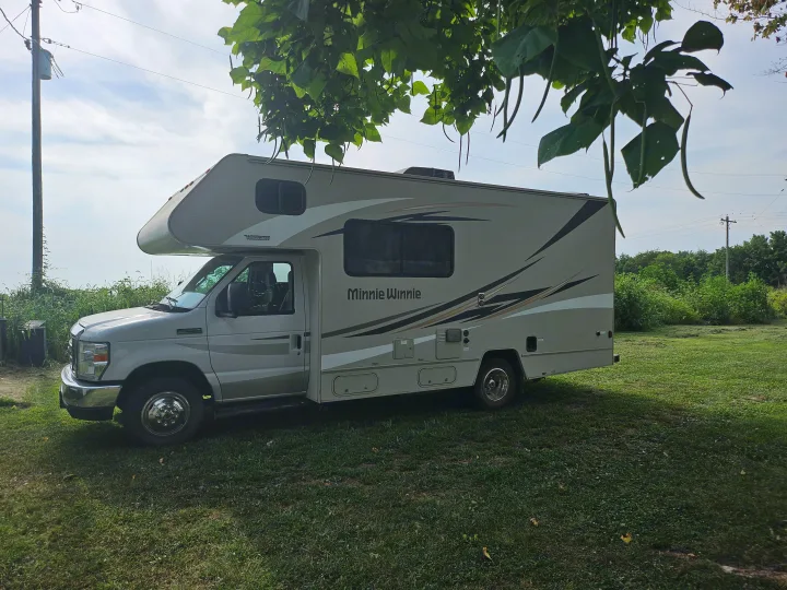 A Class C Minnie Winnie RV rental, light beige with stripes, parked on grass. Overhanging tree leaves and pods frame the scene.. Winnebago Minnie Winnie 2018