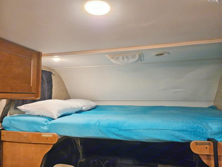 Cozy RV rental sleeping area: a bright bunk with a blue sheet, white pillow, ceiling light, and vent.. Winnebago Minnie Winnie 2018