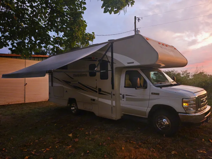 Minnie Winnie RV rental with awning extended, parked on grass. Golden hour light creates a scenic backdrop for camping.. Winnebago Minnie Winnie 2018