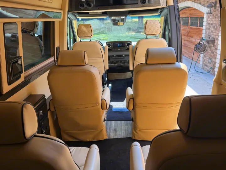 Spacious, cream-colored luxury rv rental interior with comfortable captain's chairs, looking out at a garage and trees.. Midwest Automotive Designs Luxe Cruiser 2021