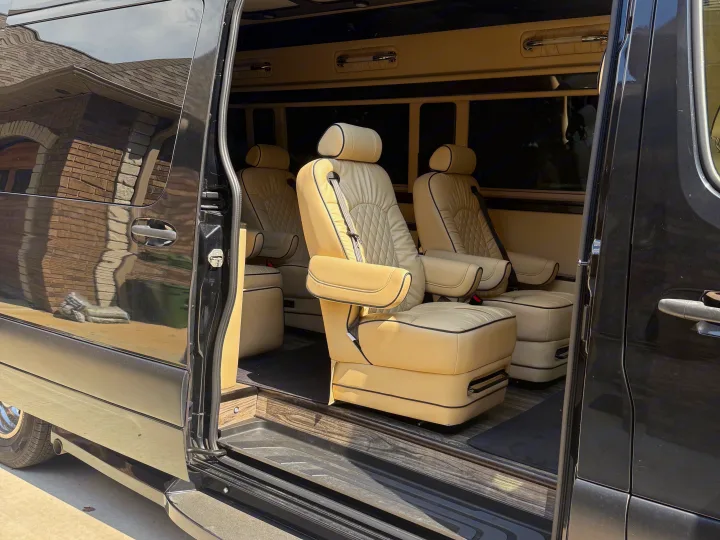 Luxurious cream leather captain's chairs inside an RV rental conversion van with a wood floor and open sliding door.. Midwest Automotive Designs Luxe Cruiser 2021