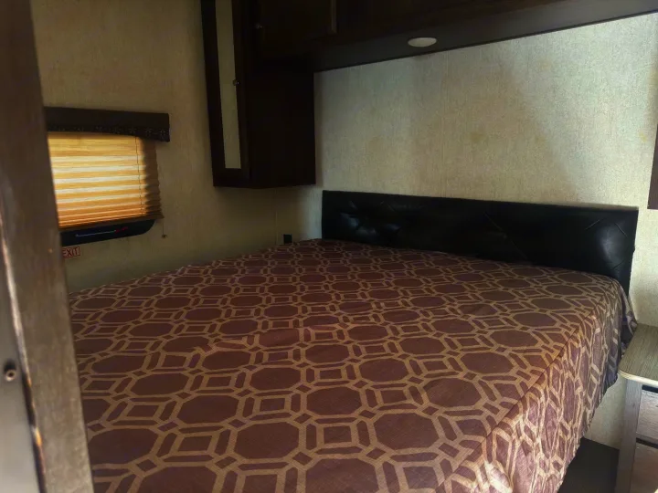 RV rental bedroom with a geometric bedspread, padded black headboard, window with blinds, and dark wood overhead cabinets.. Starcraft GPS 2018