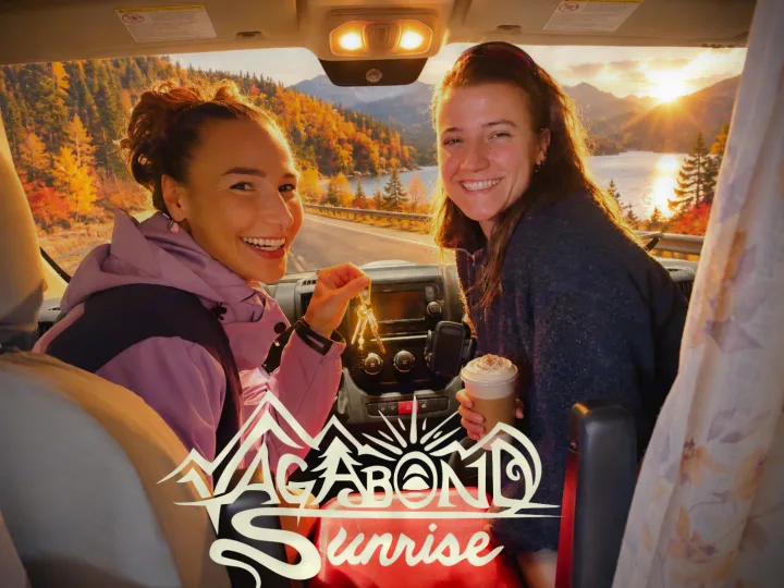 Two smiling women inside an RV rental. Driver holds keys; passenger holds coffee. Scenic autumn lake & sunrise outside.. Dodge Ram ProMaster 2500 High Top 2018