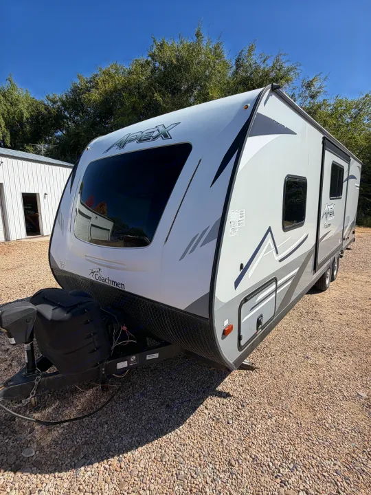 Explore the outdoors with this white & gray Coachmen Apex travel trailer, ready for rv rental adventures on gravel.. Coachmen Apex 2020