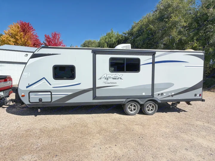 Grey APEX rv rental travel trailer with fall trees & a building in the background. Ready for your next camping trip!. Coachmen Apex 2020