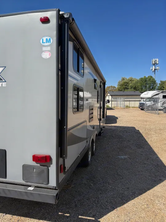 A grey RV rental travel trailer with awning, parked outdoors on a sunny gravel lot.. Coachmen Apex 2020