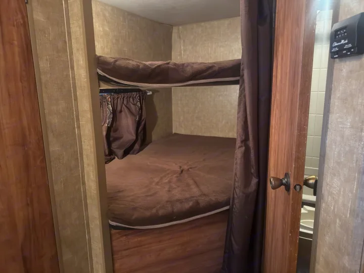 RV rental bunk beds with brown bedding and privacy curtains. A thermostat and glimpse of the bathroom are visible on the right.. KZ C291IKS 2015