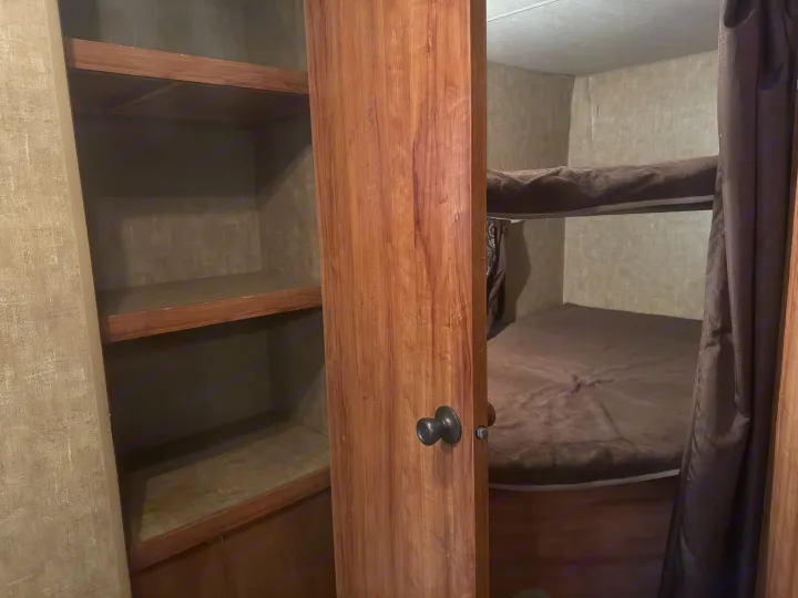 RV rental bunk bed sleeping area with dark bedding, alongside open storage shelves. Functional and compact RV interior.. KZ C291IKS 2015