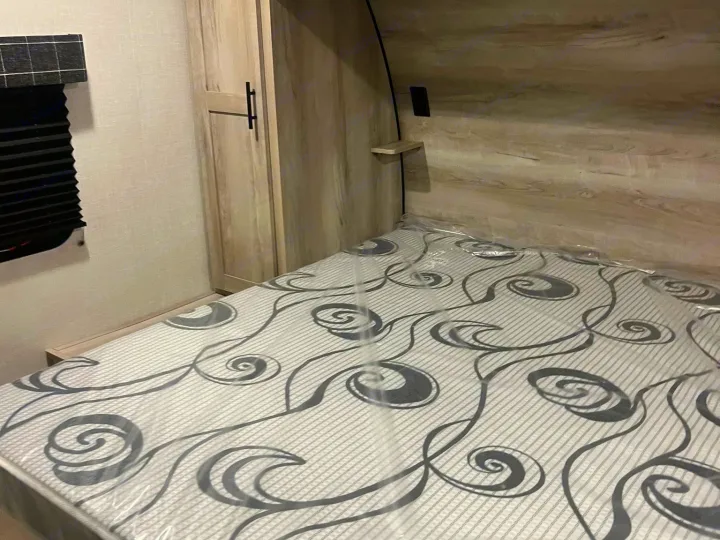 Inside a new RV rental bedroom, featuring a light-toned wood interior, wardrobe, window, and a mattress still wrapped in plastic.. Other Other 2024