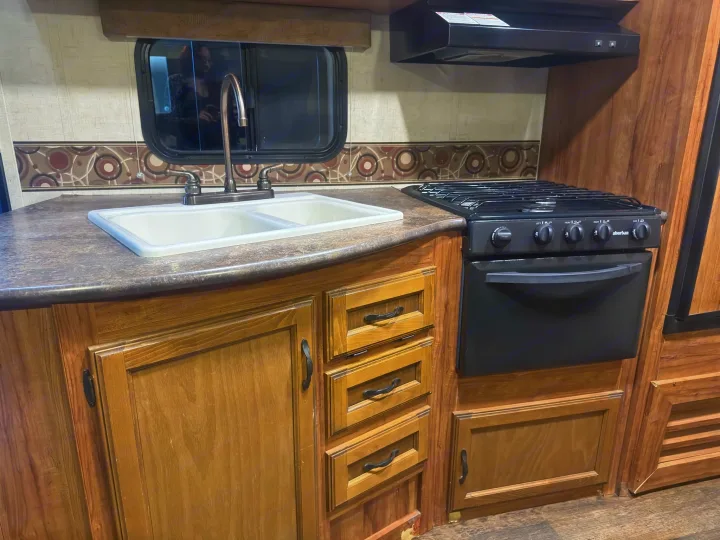 RV rental kitchen featuring a double sink, gas stove with oven, dark countertop, and wooden cabinets.. KZ C250BHS 2015