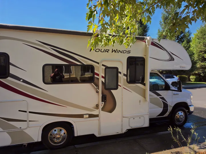 Side view of a white Four Winds RV rental, parked on a sunny day with a photographer's reflection in the window.. Other Other 2020