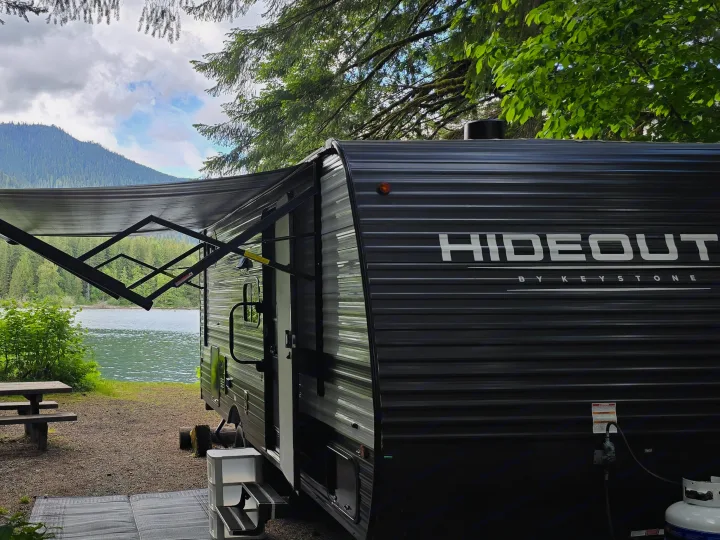 Scenic rv rental campsite by a lake and mountain, with a picnic table and extended awning. Ideal nature escape.. Keystone RV Hideout Sport 2024