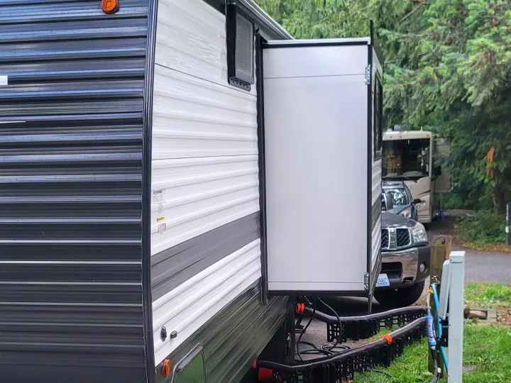 A parked RV rental with an extended slide-out and utility connections at a tree-lined campground, perfect for camping.. Keystone RV Hideout Sport 2024