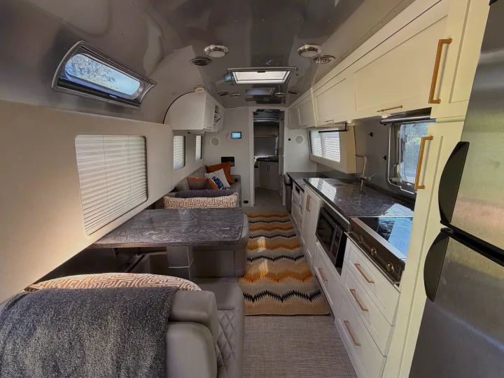 Modern rv rental interior, bright and spacious. Features a well-appointed kitchen, dining area with sofa, and windows with blinds.. Airstream Classic 2021