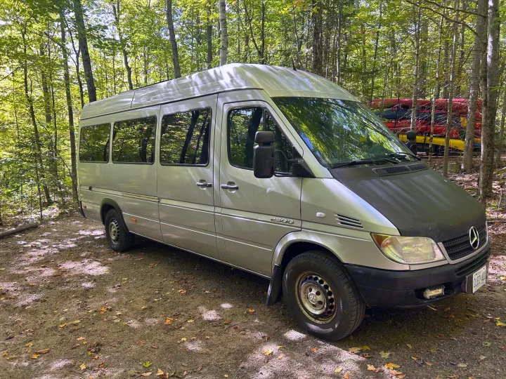 Silver Mercedes Sprinter 2500 RV rental parked in a sunlit forest, ready for outdoor adventure with kayaks.. Dodge Sprinter 2005