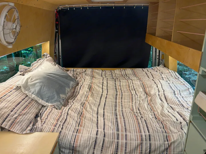 RV rental's cozy bed with striped duvet, window forest view, wall fan, and built-in storage. Ideal for road trips.. Dodge Sprinter 2005