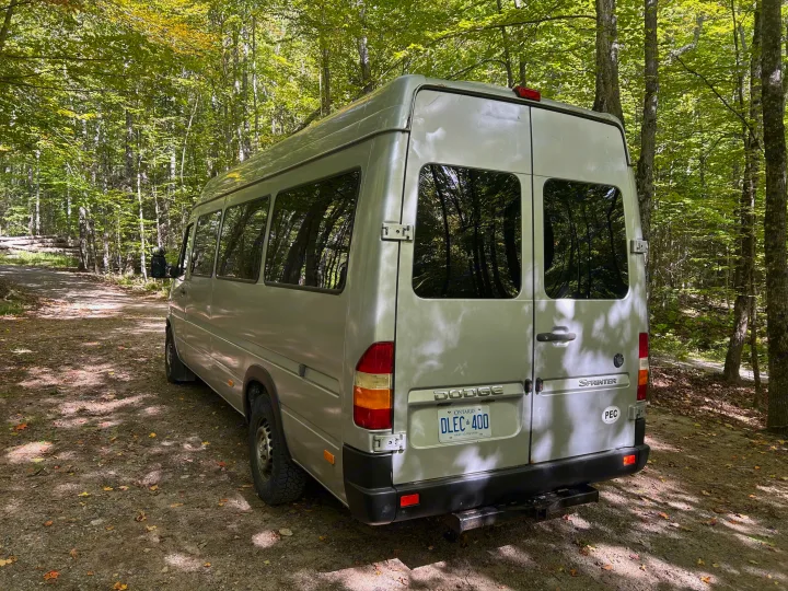 Silver Dodge Sprinter van, an RV rental, parked on a dirt forest road among trees with green and autumn leaves.. Dodge Sprinter 2005