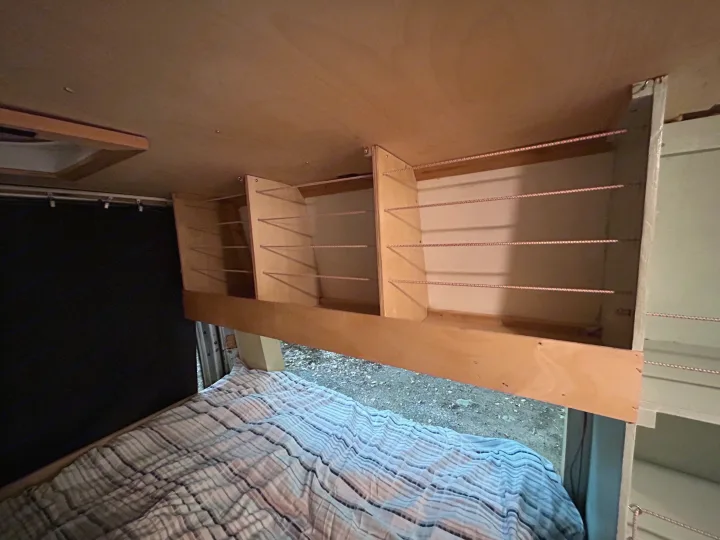 Cozy RV rental interior features custom wooden overhead storage shelves with cords, a comfortable bed, and a window view.. Dodge Sprinter 2005