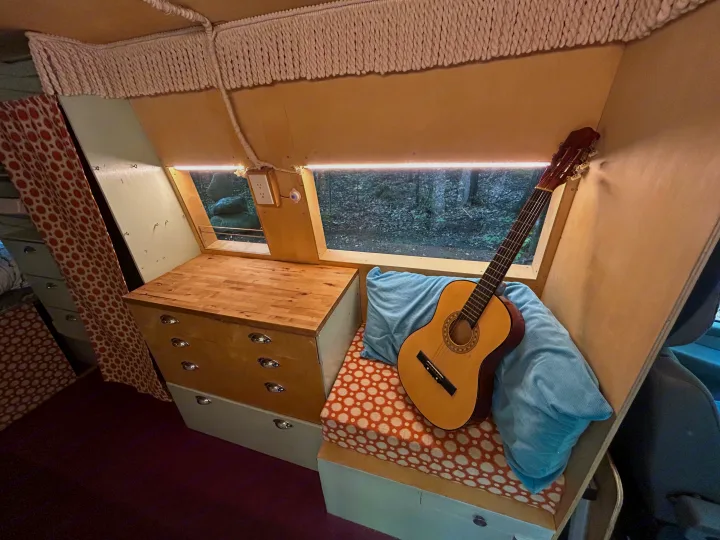 RV rental interior with a guitar, wooden drawers, and a window framing a forest view, offering a cozy travel setup.. Dodge Sprinter 2005