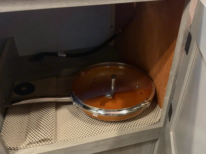 Practical RV rental kitchen storage: a copper frying pan and lid on a non-slip liner inside a cabinet with a gas line.. Sunlite 13BHLTD 2027