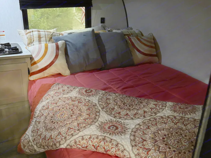 Cozy rv rental bed with colorful pillows, pink comforter, and patterned blanket. A compact stove and window view complete the interior.. Sunlite 13BHLTD 2027