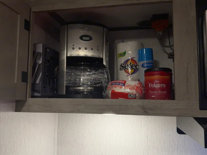 Inside an RV rental cabinet: a Gevalia coffee maker, Folgers coffee, paper towels, flatware, and cleaning spray.. Sunset Park RV Sun Lite LTD 2024