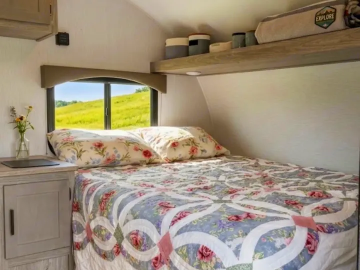 A cozy RV rental bedroom features a floral bed, window with a lush green view, and practical overhead shelving.. Sunset Park RV Sun Lite LTD 2024