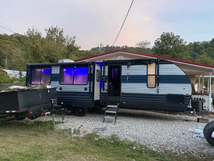Cherokee RV rental with bright blue interior lights, setup on gravel with an attached porch and hills in the background at dusk.. Forest River Cherokee 2023