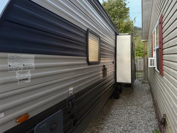 Side view of a parked RV rental with its slide-out extended, nestled close to a house with a window AC unit. Gravel path.. Forest River Cherokee 2023