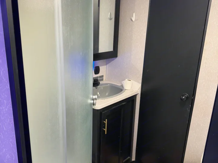 Compact rv rental bathroom featuring a frosted shower, sink with toilet paper, mirror, black cabinet, and a dark door.. Forest River Cherokee 2023
