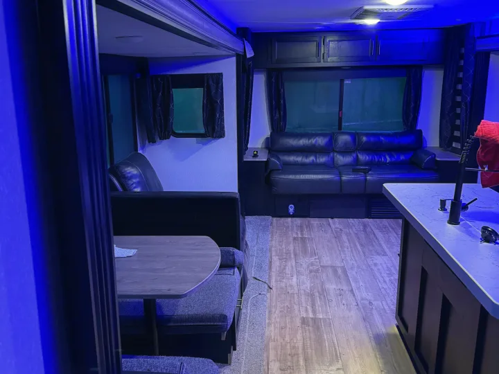 Modern RV rental interior with blue ambient light, featuring a leather sofa, dining booth, and kitchen counter. Wood flooring visible.. Forest River Cherokee 2023