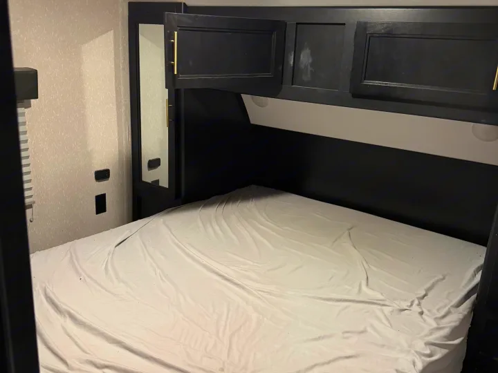 Inside an RV rental bedroom showing a bed with white sheets, overhead black cabinets, and a mirror.. Forest River Cherokee 2023