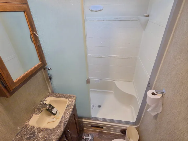 Compact RV rental bathroom with sink, mirror, toilet, and a shower stall featuring a frosted glass door. Clean and ready for your trip.. Keystone RV Laredo 2016